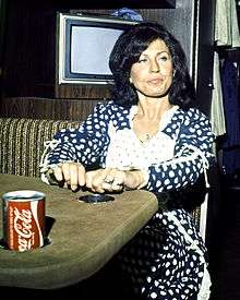 A black-haired woman in a dark blue outfit with white polka dots, looking contemplative while sitting at a table