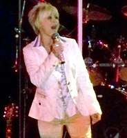A blonde woman wearing a white jacket singing into a microphone