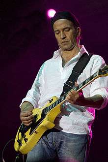 A full color picture of Urquijo holding a yellow electric guitar.  He wears a black cap, white shirt, and blue jeans.