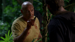 A picture of John Locke, a white man who is approximately 48 years old, pointing at a moth cocoon with a knife, while Charlie Pace watches