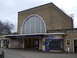 A beige-bricked building with a dark blue, rectangular sign reading "LOUGHTON STATION" in white letters all under a blue sky with white clouds