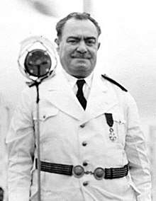 Half-length photograph of a middle-aged, man in a white uniform, standing before a microphone