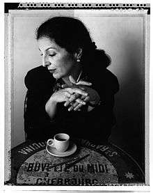 Black and white image of Fili sitting at table. Her hands are crossed over a cup of coffee and the table is decorated with Modernist type.
