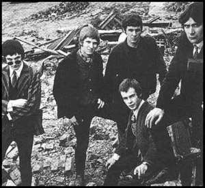 Five men at a demolished house site. Man at left stands, arms folded across chest and peers over sunglasses. Second man has left leg propped on building material. Third man is partly obscured by fourth man sitting in front with hands on knees. Fifth man has right leg propped with his arm on knee and is partly out of shot.