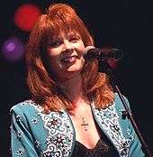 A woman with long red hair wearing a light blue jacket, standing at a microphone