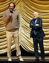 Two middle-aged Caucasian men stand before a yellow curtain on stage.
