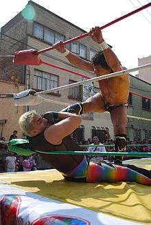A Mexican wrestler placing his foot on the throat of a seated opponent.
