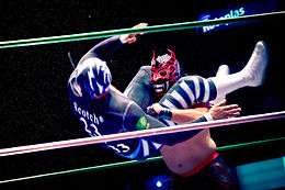A color photograph of a wrestler in black and white clothes, including a black and white mask executing a head scissors takedown on a wrestler in black and red mask with ornamental horns.