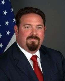 A photograph of a man with a goatee looking at the viewer while wearing a navy suit jacket, a white dress shirt, and a red necktie