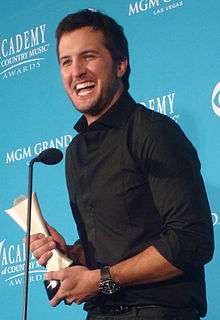A young man with dark hair wearing a black shirt, speaking into a microphone and holding a trophy