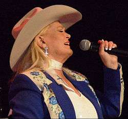 A blonde woman wearing a cowboy hat and a blue jacket with an elaborate floral pattern, singing into a microphone