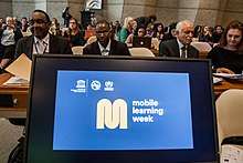 A computer screen at the front of a room of policy makers shows the Mobile Learning Week logo