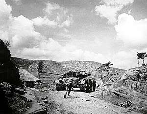 A tank advances up a hill followed by men in military uniform