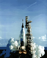 A rocket firing just above its launch pad, next to a metal structure, with a large cloud of smoke at its base and some smoke around the upper section of the rocket. Flame can be seen below the cloud.