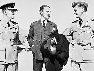 Mustachioed man in business suit flanked by two men in military uniforms