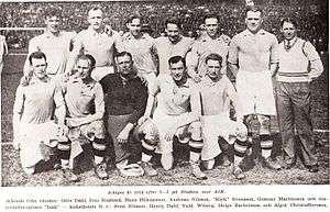 A black-and-white photograph of a football team lining up after a match. Six players wearing light shirts, white shorts and dark socks are standing behind a row of five kneeling players with the same apparel except for the man in the middle of the row of kneeling players who is wearing a dark shirt, dark shorts and dark socks. A man with casual clothing is standing to the right side of the row of standing players.