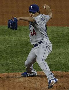 A man wearing a gray baseball uniform and blue baseball cap throws a baseball with his right hand.