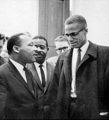 Malcolm X and Martin Luther King speak to each other thoughtfully as others look on