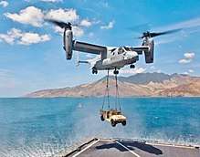 A MV-22 Osprey with its rotors up to vertical with a HMMWV vehicle hanging by two sling wires.