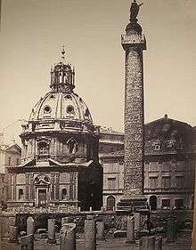 A photograph of the Forum of Trajan in Rome. In the foreground are a number of small broken stone columns. In the background on the left is the church of Santa Maria di Loreto, while on the right is the Column of Trajan.