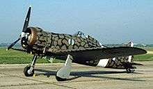 a single-engine monoplane with green and black mottled camouflage paint parked on the tarmac