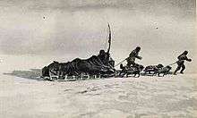 A loaded sledge being pulled across an icy surface by two figures and a team of dogs