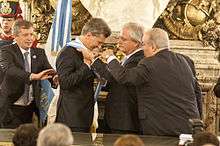 A smiling Macri puts on a blue-and-white sash with the help of Federico Pinedo and two other men