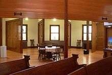 Pews in foreground; looking under half-closed tambour doors to room divided by radial partitions hung with folding doors