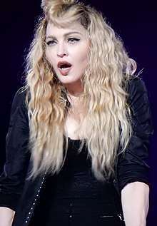 Madonna in a black dress and jacket and long blond hair singing.