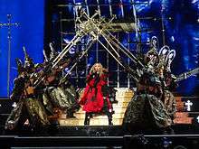 Madonna in a red kimono-like dress in the middle of the stage, flanked by dancers in Samurai costumes holding up giant crosses above her.