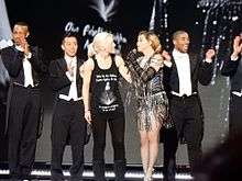 Madonna standing onstage speaking with an audience member, flanked by her dancers in coattails.