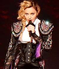 Madonna playing an ukulele and smiling and singing, while looking to her right.
