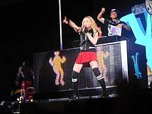 Madonna dancing on a DJ's desk wearing red shorts and a black sleeveless shirt. She's holding on to a pole with her left hand and holds a microphone to her mouth with her right hand.