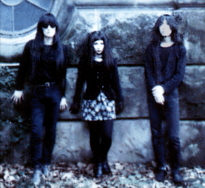 A full body bluescale photography of two men and a woman between them. All of them are standing while leaning their backs on a wall made of retangular block rocks. They are straight looking at the camera. From left to right: The first person is a man who has about the same height as the other man; he's white; has long black hair with bangs; is using dark clothes and dark sunglasses. The second person is a woman; she's smaller than the other two people; she's white; has long, black hair with bangs; she's using back gloves; her clothes are dark, but her skirt, which has light and dark tones The third person is a man who has the same height as the other man; he's white; his black hair leans on his shoulders; he's wearing dark clothes; he tightly hold his hands down.