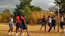 Malawian young women playing netball on a dirt court. One player is preparing to shoot for goal, while other players in and around the shooting circle look on. Bibs are not worn by any of the players.