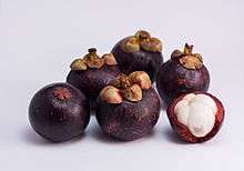 Photograph of several purple mangosteen fruits. One has been partially peeled revealing white flesh divided into five sections.