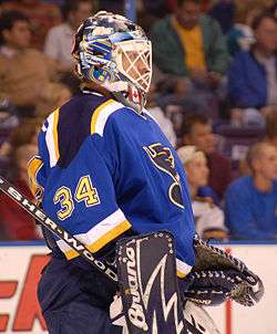 Hockey player in blue uniform with the number 34 on it and goaltender's gear. He faces forward.