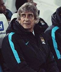 A photograph of a gentleman in his 60s. He is sitting on a red side bench. He is wearing a navy blue hooded jacket with a Manchester City crest on the left breast and a white logo of the Nike sponsor on the right breast.