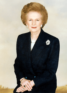 portrait at half length of an older white woman with coiffed, light golden brown hair, wearing jewellery, dressed in a dark suit, hands crossed, against a cloudy backdrop