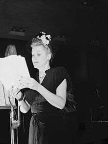 A blonde woman with flowers in her hair, looking at some papers which she is holding in her hands