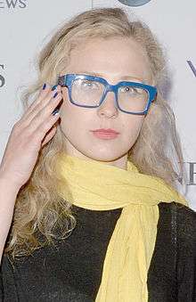 A bleach blonde woman with bright blue glasses and a yellow scarf.