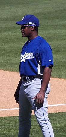 A dark-skinned man in a blue baseball jersey and cap and gray baseball pants wearing sunglasses and standing on a baseball field