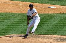 Mariano Rivera in a white pinstriped baseball uniform and navy blue cap stands on a dirt mound. He is striding forward to the right as he clutches a baseball.