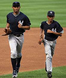 Mariano Rivera and his son wearing navy blue hats and baseball jerseys as they jog on a baseball field.