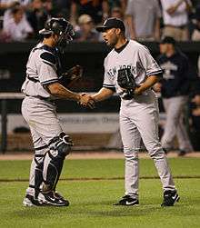 Jorge Posada wearing catcher's equipment shakes hands with Mariano Rivera on a grass field.