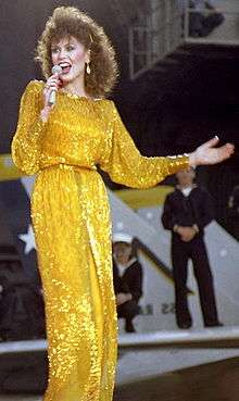 A young woman with long brown hair, wearing a long yellow dress, singing into a microphone