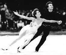 An ice dance couple performing a routine. The man, on the right, is dressed with a dark suit and holds his white-dressed partner by her waist and left hand.