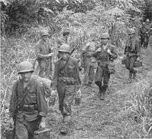 Black and white photo of four men wearing military uniforms and helmets carrying bags and boxes along a trail. Several other men are standing at the edge of the trail, and behind them.
