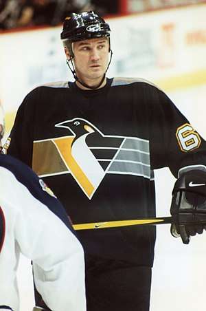 Upper body of a man wearing a black uniform with a stylized penguin in yellow, grey and white on the chest. He is looking into the distance.