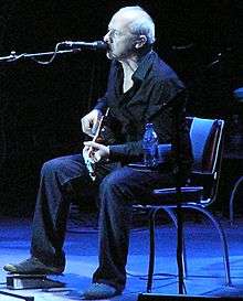 Image of Mark Knopfler sitting in a chair onstage playing guitar and singing